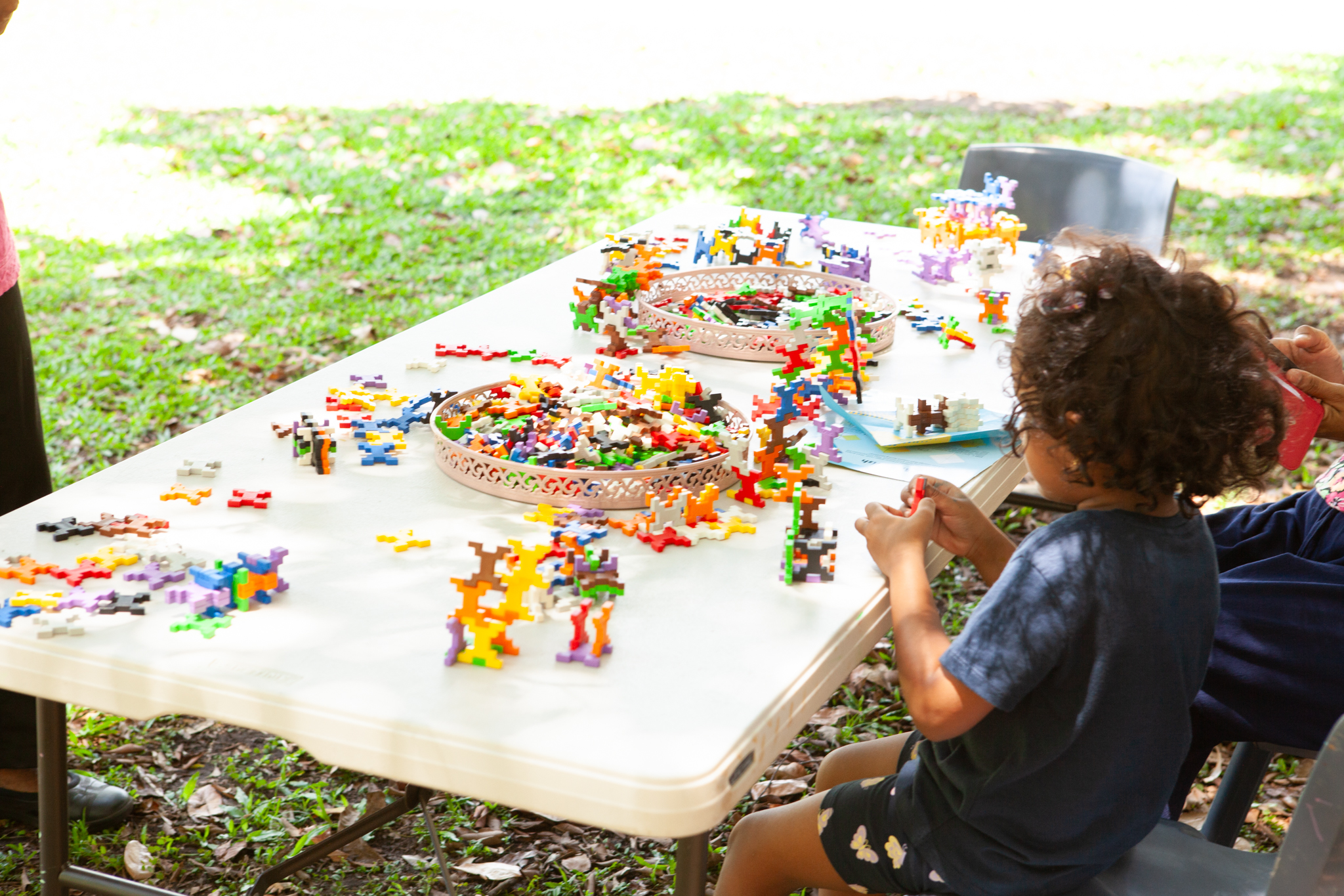 A young child sits at an outdoor table playing with colourful interlocking building blocks. The table is covered with various pieces and structures, and baskets hold additional blocks. The setting appears to be a grassy park area, suggesting a playful and engaging outdoor activity.