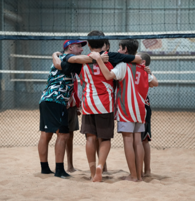 group of young people in matching red jerseys standing in a huddled circle on the sand of a volleyball court