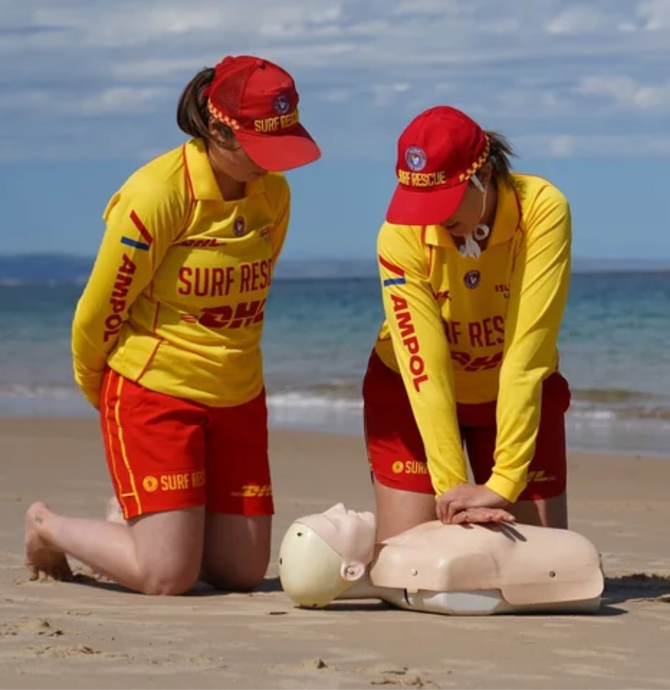 two girls in yellow and red surf life saving beach outfits performing CPR on a dummy