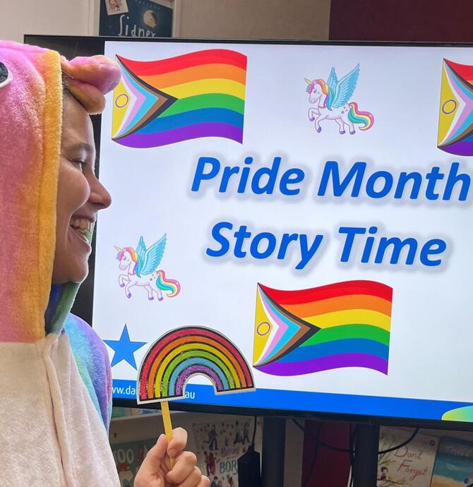A person dressed as a unicorn gazing at a colourful sign filled with rainbows that reads “Pride Month Story Time.”