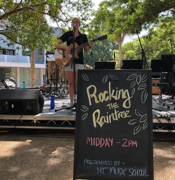 young person playing guitar in front of sign that says Rocking the Raintree